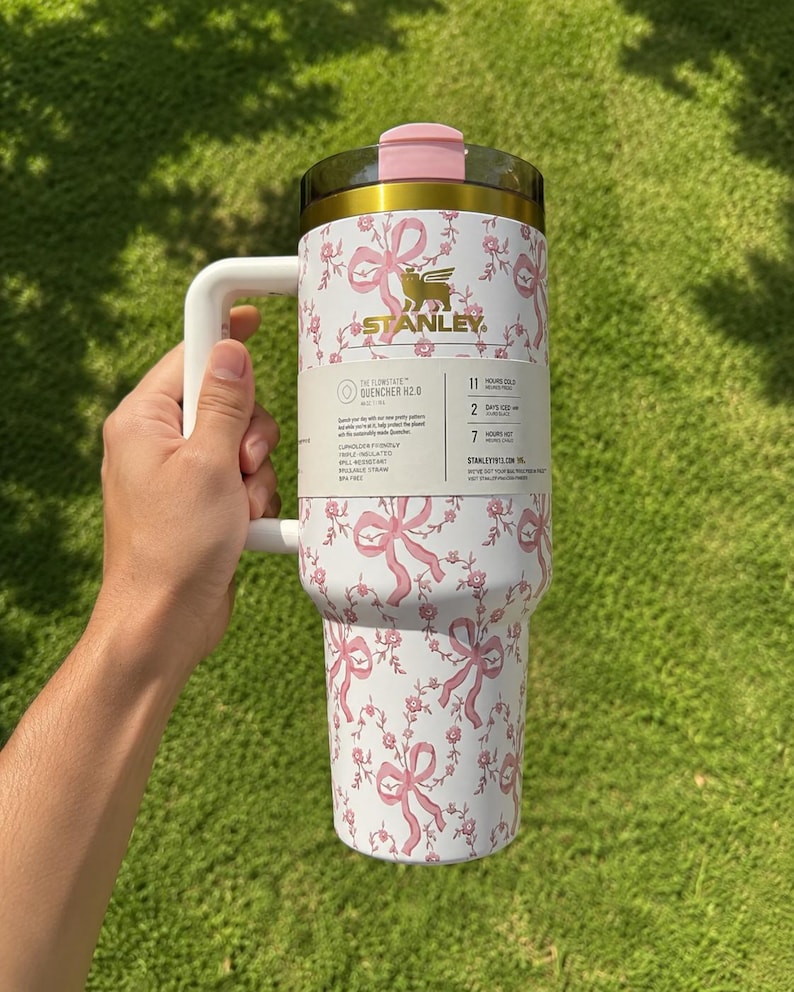 May include: A white Stanley Quencher H2.0 Flowstate tumbler with a pink bow and floral pattern. The tumbler has a handle, a pink lid, and gold accents. The Stanley logo is visible. The tumbler is designed to keep drinks cold for 11 hours.
