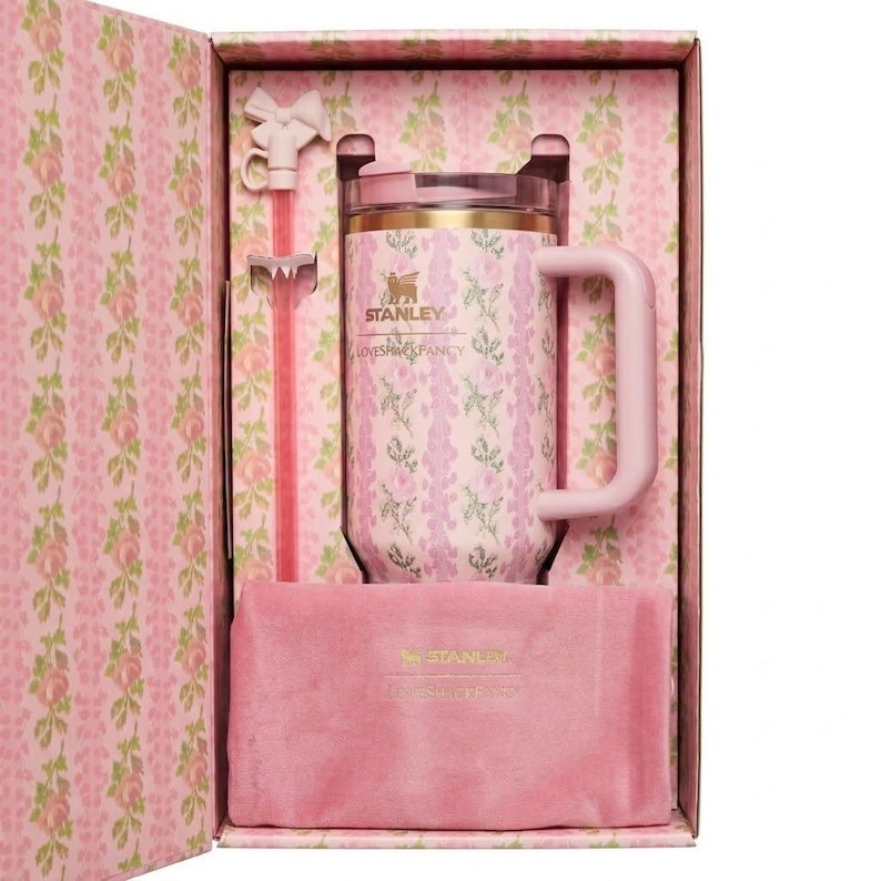 May include: A pink Stanley mug with a floral pattern, presented in a matching gift box. The mug has a handle and a lid, and includes a pink straw and a pink velvet pouch. The box and mug feature the "Stanley" logo.