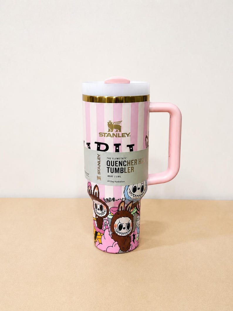 May include: A pink and white striped Stanley Quencher H2.0 Flowstate tumbler with a pink handle and lid. The tumbler features a colorful cartoon design and holds 40 oz (1.18L) of liquid.