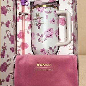 Op de afbeelding: Een witte Stanley-mok met een roze rozenpatroon, een roze fluwelen etui en een paarse rietje met een strik, alles in een bloemenprint doos. De mok heeft een handvat en de Stanley en Lovesackfancy logo's.