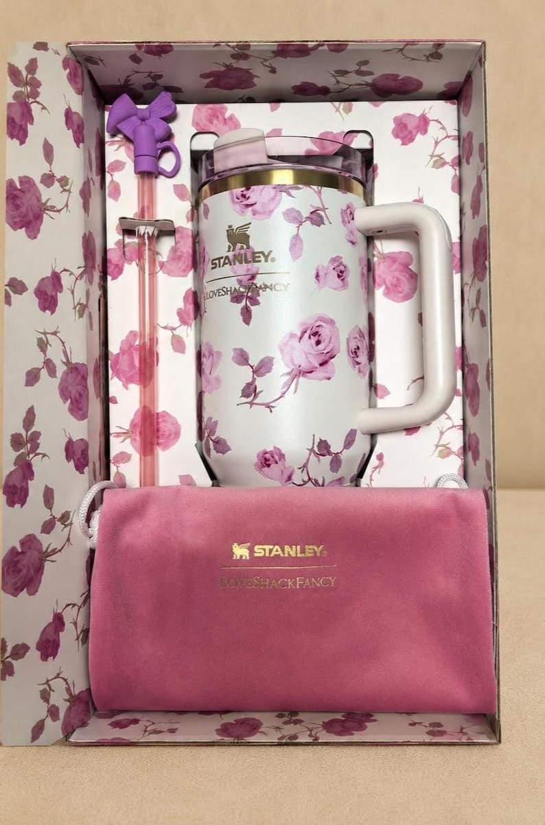 Op de afbeelding: Een witte Stanley-mok met een roze rozenpatroon, een roze fluwelen etui en een paarse rietje met een strik, alles in een bloemenprint doos. De mok heeft een handvat en de Stanley en Lovesackfancy logo's.