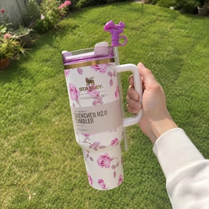 Op de afbeelding: Witte en roze bloemen Stanley Quencher H2.0 Tumbler met een handvat en een paarse strikvormige strooitopper. De tumbler heeft de tekst "Stanley" en "Loveshafancy" erop.