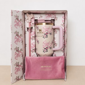 Pode incluir: Um conjunto de presente de caneca de viagem Stanley em uma caixa com estampa floral. A caneca isolada tem um fundo creme com laços e rosas cor-de-rosa. O conjunto inclui um canudo rosa, uma bolsa de veludo rosa e uma alça rosa.