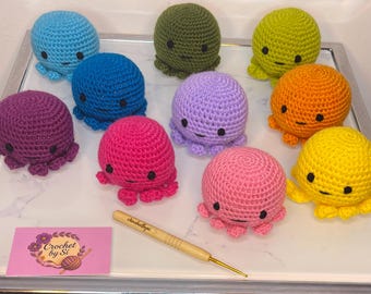 May include: A selection of colourful, crocheted octopus toys. The octopuses are in shades of blue, green, purple, pink, orange, and yellow. Each has a round head, black eyes, and eight tentacles. A crochet hook and business card are also visible.