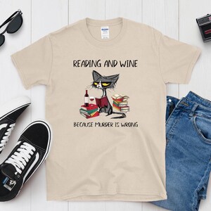 May include: Beige t-shirt with the text "READING AND WINE BECAUSE MURDER IS WRONG" and a cartoon cat reading a book with a wine bottle and glass. Black sunglasses, black and white sneakers, blue jeans, and a vintage camera are also in the image.