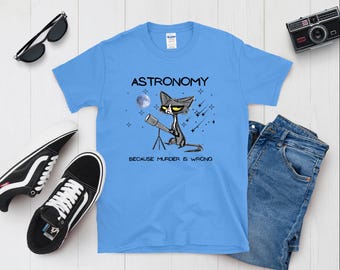May include: A light blue t-shirt featuring a cartoon cat looking through a telescope. The text on the shirt reads "Astronomy because murder is wrong."
