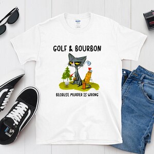 May include: White t-shirt with a cartoon cat holding a golf club and a bottle of bourbon. The text on the shirt reads "Golf & Bourbon Because Murder is Wrong."