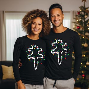 May include: Two people wearing black long-sleeved shirts with a white silhouette of a person tangled in Christmas lights. The lights are red, green, yellow, and blue.