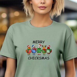 May include: Sage green t-shirt featuring the text "MERRY CHICKSMAS" above a graphic of five cartoon chickens wearing Christmas sweaters and Santa hats. The chickens are in various colors, including red, green, and blue.