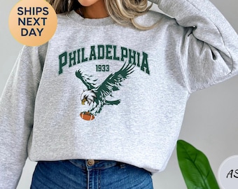 Könnte beinhalten: Hellgraues Sweatshirt mit dem grünen Schriftzug "PHILADELPHIA 1933" und einer Grafik eines Adlers, der einen Football hält. Das Sweatshirt hat einen Rundhalsausschnitt.