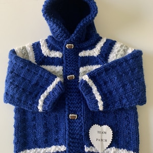 May include: A blue knitted hooded jacket with white stripes on the sleeves and a white stripe along the bottom. The jacket has football-shaped buttons and a "TEAM PATCH" label. The hood has a white and blue pom-pom.