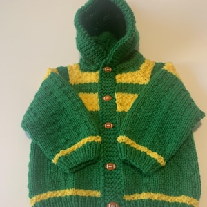 May include: Green and yellow knitted hooded sweater with football-shaped buttons. The sweater features yellow stripes on the sleeves and chest. The hood is a deep green color, and the buttons are brown with white laces.
