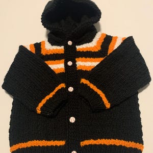 May include: Black knitted hooded cardigan with orange and white stripes, button closure, and baseball-themed buttons.