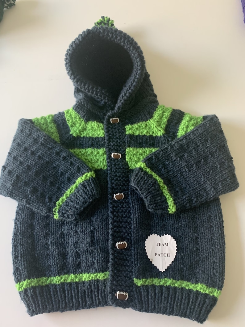 May include: A dark blue knitted baby cardigan with green stripes and football-shaped buttons. The cardigan has a hood and a small white heart-shaped tag that says "Team Patch".