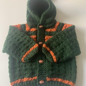 May include: A green knitted baby cardigan with orange stripes and football-shaped buttons. The cardigan has a hood with a pom-pom on top.