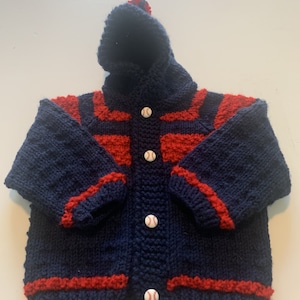 May include: A blue knitted hooded cardigan with red stripes and baseball buttons. The cardigan has a hood with a small pom-pom on top.