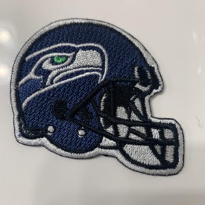 May include: A blue and white embroidered patch of a football helmet with a bird logo. The helmet is white with a black face mask and the bird has a green eye.
