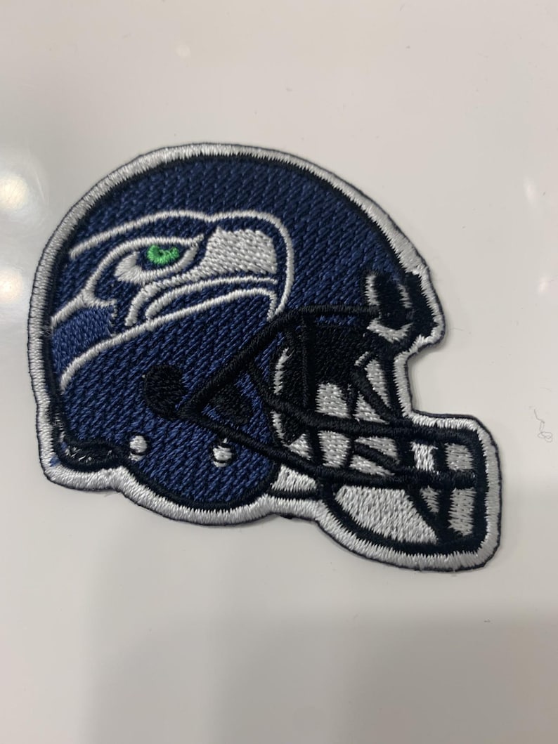 May include: A blue and white embroidered patch of a football helmet with a bird logo. The helmet is white with a black face mask and the bird has a green eye.