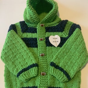 May include: Green and navy blue knitted hooded cardigan with football buttons. The cardigan has a white heart-shaped patch that says "Team Patch".