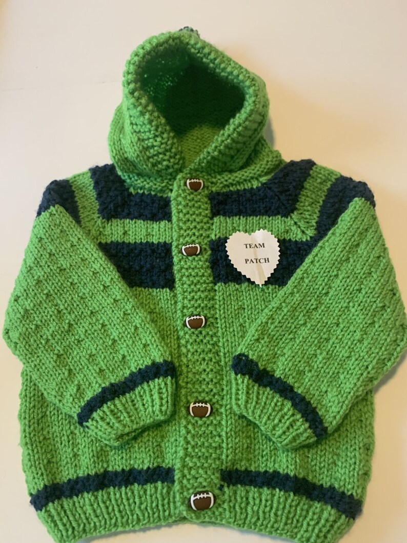 May include: Green and navy blue knitted hooded cardigan with football buttons. The cardigan has a white heart-shaped patch that says "Team Patch".