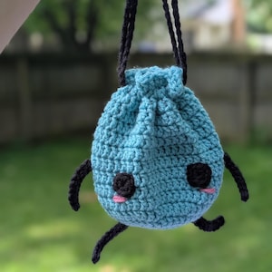 May include: A light blue crochet drawstring pouch featuring a round body with two black circular eyes and small pink stitched cheeks. The pouch has four thin black crocheted limbs and a long black crocheted handle for carrying or hanging.