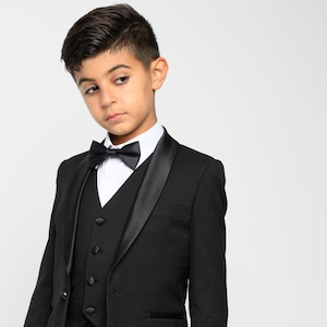 May include: A young boy wearing a black tuxedo with a satin lapel and a black bow tie. He is standing in front of a white background.