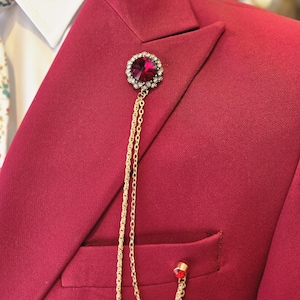 Może przedstawiać: Zbliżenie bordowej marynarki garnituru z białą koszulą i kwiatowym krawatem. Ozdobna broszka z dużym, czerwonym kamieniem szlachetnym i złotym łańcuszkiem zwisa z klapy. Marynarka ma kieszeń z małym złotym akcentem.