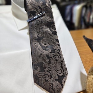 Peut inclure: Une cravate noire avec un motif cachemire marron et une pince à cravate argentée, posée sur une chemise blanche. La pince à cravate a une petite pierre transparente. L'arrière-plan est flou.