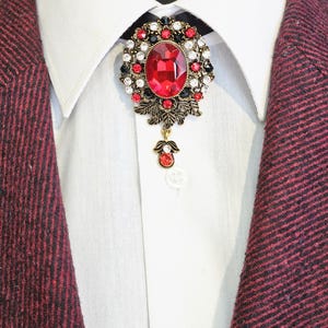 May include: A close-up of a decorative brooch with a large red oval gemstone at the center, surrounded by smaller red, black, and white stones. The brooch is set against a white shirt and a burgundy blazer.
