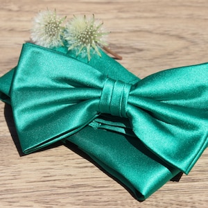 May include: A vibrant emerald green bow tie and matching pocket square set. The bow tie is pre-tied and features a satin finish. Two small, dried, white flowers are placed on top of the bow tie. The set is displayed on a wooden surface.