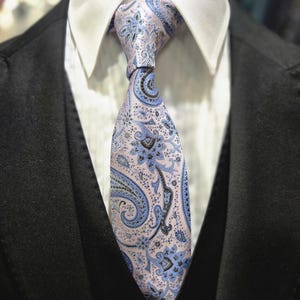 Puede incluir: Primer plano de una corbata con estampado de cachemira en tonos rosa claro, azul y negro. La corbata está anudada y se lleva con una camisa de cuello blanco y una chaqueta de traje negra. El diseño de la corbata presenta un motivo floral y de gota.