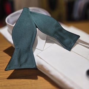 May include: A dark green bow tie made of satin fabric. The bow tie is folded and placed on a white dress shirt.