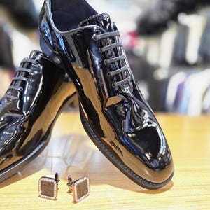 Op de afbeelding: Een paar glanzende zwarte leren schoenen met bijpassende veters. De schoenen staan naast een paar vierkante manchetknopen met een donker houten nerfcentrum en een rand van kleine, heldere stenen. Op een licht houten oppervlak.