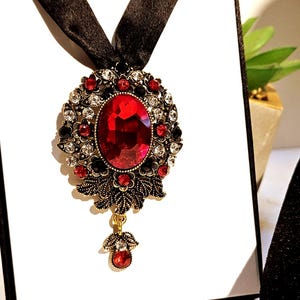 May include: A red and gold statement necklace with a large red gemstone pendant surrounded by smaller clear and black gemstones. The necklace is on a black ribbon.