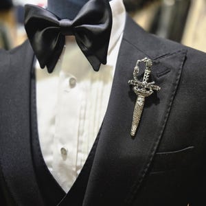 May include: A black tuxedo jacket with a matching vest and a white dress shirt. A large black bow tie is at the neck. A decorative silver sword pin is on the lapel, adding a touch of elegance to the formal attire.