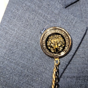 Pode incluir: Um broche de lapela dourado com um design de cabeça de leão em esmalte preto. O broche tem uma corrente anexa.