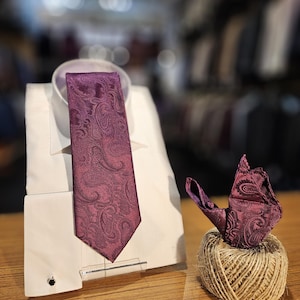 May include: A purple paisley patterned tie and pocket square set. The tie is displayed on a white shirt collar and the pocket square is folded and placed on a ball of twine.
