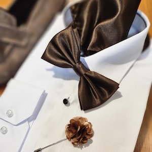 Pode incluir: Uma camisa branca com uma gravata borboleta marrom e um lenço de bolso marrom. Um broche de lapela de flor marrom está preso à camisa.