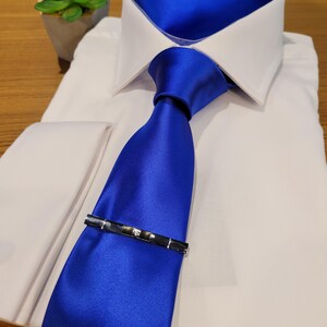 Puede incluir: Una camisa blanca con una corbata de seda azul y un pañuelo de bolsillo de seda azul. La corbata tiene un clip de corbata plateado.