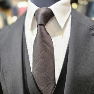 May include: A close-up of a dark brown plaid tie knotted and worn with a white collared shirt, black vest, and black suit jacket. The tie has a subtle pattern and is the focal point of the image.