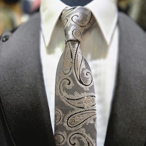 Może przedstawiać: Wzorzysty krawat z wzorem paisley w odcieniach srebra, czerni i złota jest zawiązany i noszony z białą koszulą z kołnierzykiem i czarną marynarką. Złożony wzór krawata jest punktem centralnym.