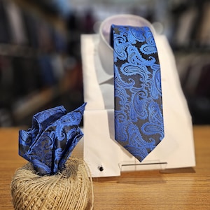 May include: A blue and black paisley patterned tie and matching pocket square. The tie is on a white shirt collar and the pocket square is on a brown twine ball.