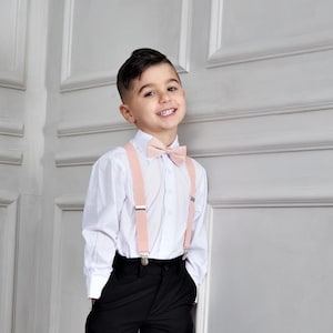 May include: A young boy wearing a white button-down shirt with pink braces and a pink bow tie. He is standing in front of a white wall with wainscoting.