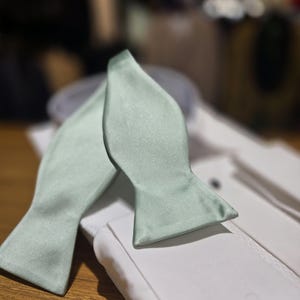 May include: A sage green bow tie made of satin fabric. The bow tie is folded and placed on a white shirt.