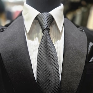 May include: A close-up of a formal outfit featuring a black suit jacket, a white dress shirt, and a gray tie with a striped pattern. The tie is knotted, and the suit jacket has a satin lapel. The overall look is classic and sophisticated.