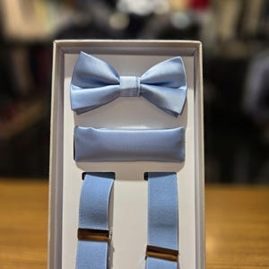 May include: A light blue formal set including a bow tie, pocket square, and suspenders. The bow tie is pre-tied. The suspenders have gold-colored metal clasps. The set is presented in a white box, ideal for gifting or special occasions.