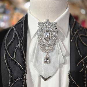 Puede incluir: Una camisa blanca formal con una broche decorativa de plata y pedrería. El broche es el punto focal, con una gran piedra central rectangular y detalles intrincados. La chaqueta negra tiene adornos de cuentas.