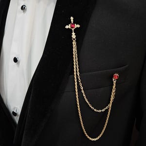 May include: A gold-toned cross with a red gemstone pendant hangs from a double chain. The chain is attached to a black suit jacket.
