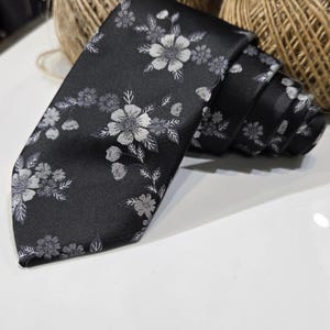 May include: A black necktie featuring a repeating pattern of silver-grey flowers and leaves. The fabric has a smooth, satin-like finish, and the tie is coiled at the end, displaying the intricate floral embroidery across the entire surface.