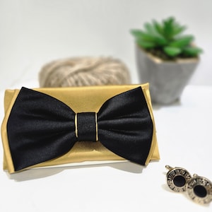 May include: A black satin bow tie with a gold satin backing. The bow tie is centered on a gold rectangle. Two gold and black cufflinks are in the lower right corner. A small succulent plant and a ball of twine are in the background.